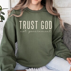 以下が含まれることがあります： 白い文字で「TRUST GOD not government」と書かれた緑色のスウェットシャツ
