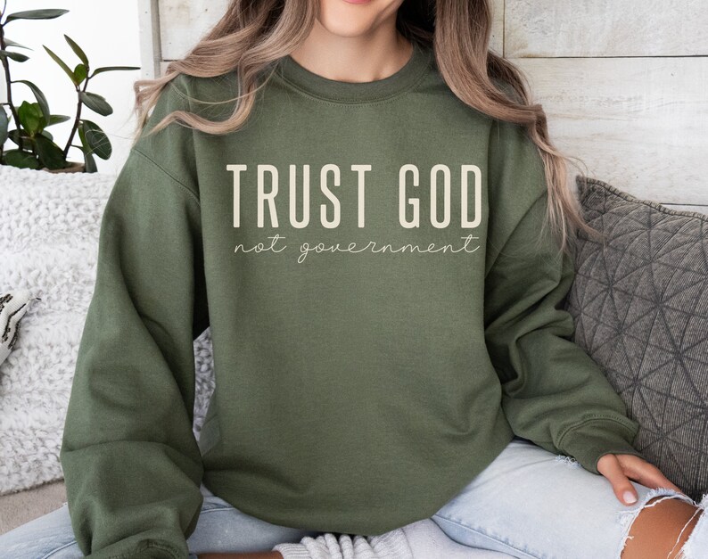 以下が含まれることがあります： 白い文字で「TRUST GOD not government」と書かれた緑色のスウェットシャツ