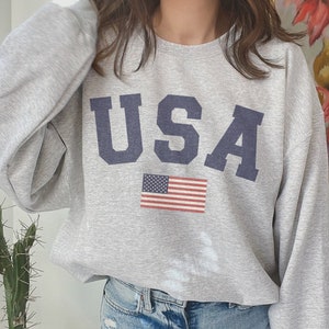 Op de afbeelding: Een grijs sweatshirt met het woord "USA" in blauw op de voorkant. Een kleine Amerikaanse vlag is onder het woord gedrukt.