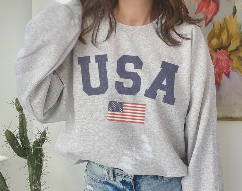 Op de afbeelding: Een grijs sweatshirt met het woord "USA" in blauw op de voorkant. Een kleine Amerikaanse vlag is onder het woord gedrukt.