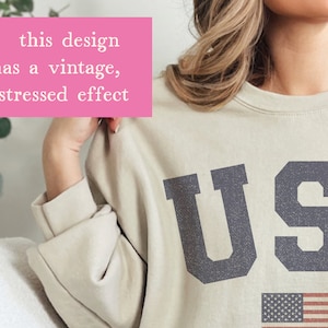 Op de afbeelding: Een beige sweatshirt met een vintage, verweerd effect met de tekst "USA" in donkerblauw met een kleine Amerikaanse vlag eronder.