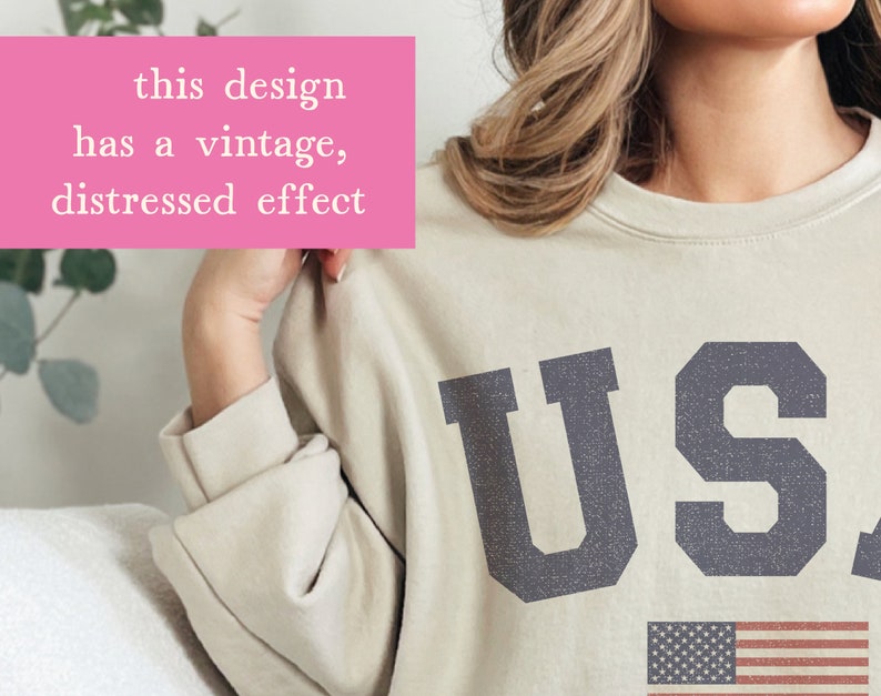 Op de afbeelding: Een beige sweatshirt met een vintage, verweerd effect met de tekst "USA" in donkerblauw met een kleine Amerikaanse vlag eronder.