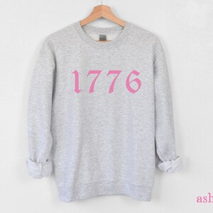 May include: A gray sweatshirt with a pink 1776 printed on the front.