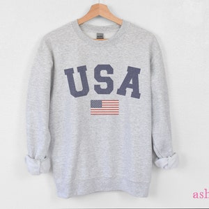Op de afbeelding: Een grijs sweatshirt met het woord "USA" bedrukt in donkerblauwe letters en een kleine Amerikaanse vlag eronder.