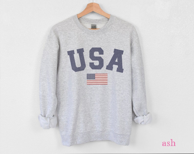 Op de afbeelding: Een grijs sweatshirt met het woord "USA" bedrukt in donkerblauwe letters en een kleine Amerikaanse vlag eronder.