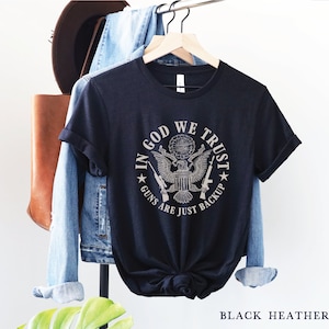 May include: Black heather t-shirt with a graphic print of the Great Seal of the United States. The graphic includes the text "In God We Trust" and "Guns Are Just Backup".