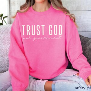以下が含まれることがあります： 白文字で「TRUST GOD not government」と書かれたピンク色のスウェットシャツです。
