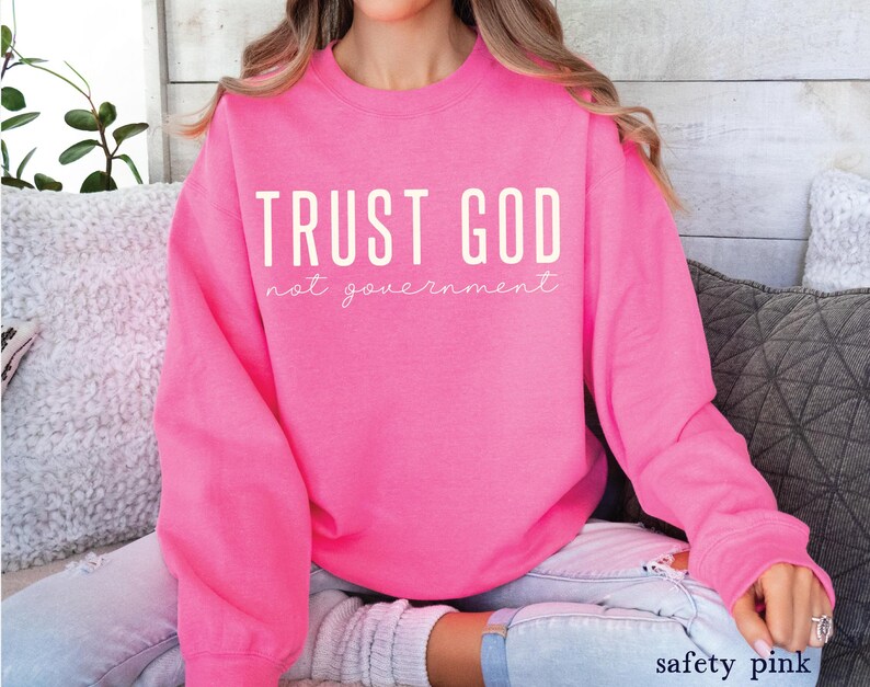 以下が含まれることがあります： 白文字で「TRUST GOD not government」と書かれたピンク色のスウェットシャツです。