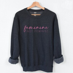 K&ouml;nnte beinhalten: Dunkelgrauer Pullover mit dem Text "feminine not feminist" in rosa Schrift.