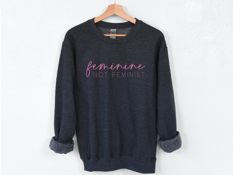K&ouml;nnte beinhalten: Dunkelgrauer Pullover mit dem Text "feminine not feminist" in rosa Schrift.