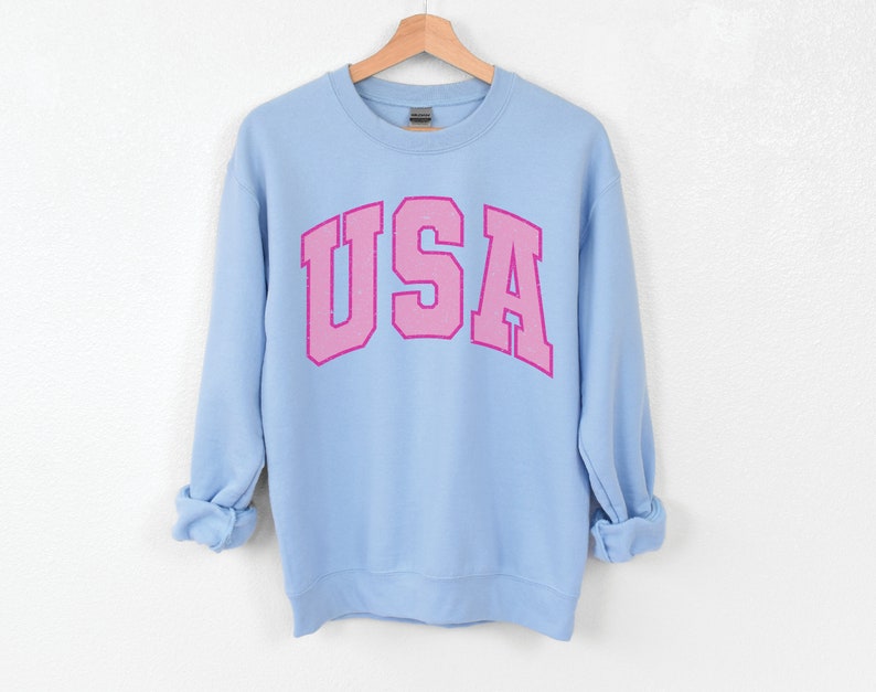 以下が含まれることがあります： ピンク色の「USA」の文字がアーチ状にデザインされた、水色のスウェットシャツ。白い背景に木製のハンガーに掛けられています。袖は折り返されています。