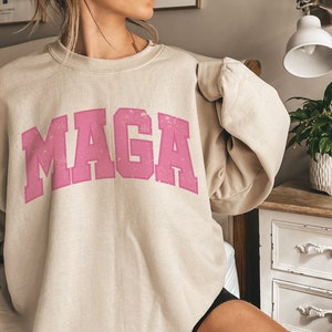 May include: A beige sweatshirt with a pink "MAGA" graphic printed on the front. The sweatshirt is made of a soft, comfortable material and has a relaxed fit.