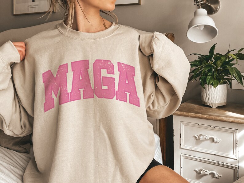 May include: A beige sweatshirt with a pink "MAGA" graphic printed on the front. The sweatshirt is made of a soft, comfortable material and has a relaxed fit.