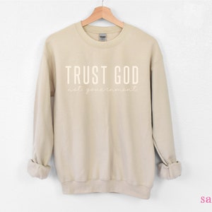 以下が含まれることがあります： サンドカラーのトレーナーに「TRUST GOD not your government」という白い文字がプリントされています。