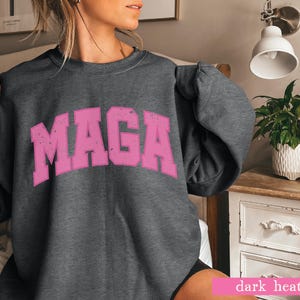 May include: A dark heather grey sweatshirt with the word "MAGA" printed in pink on the front.