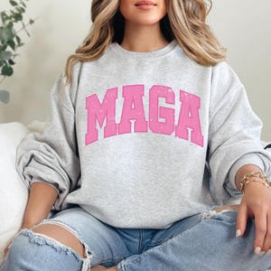 May include: A gray sweatshirt with a pink "MAGA" graphic printed on the front. The sweatshirt is worn by a person with long blonde hair.