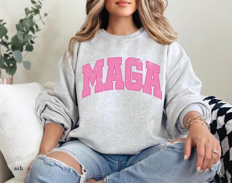 May include: A gray sweatshirt with a pink "MAGA" graphic printed on the front. The sweatshirt is worn by a person with long blonde hair.