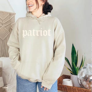 May include: A sand-colored hoodie with the word "patriot" printed in a gothic font across the chest. The hoodie has a drawstring hood and a front pocket. The model is wearing blue jeans.