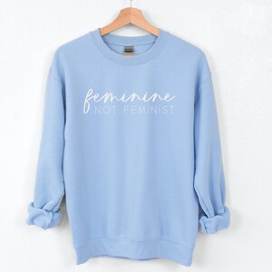 K&ouml;nnte beinhalten: Ein hellblauer Pullover mit dem Text "feminine not feminist" in wei&szlig;en Buchstaben.
