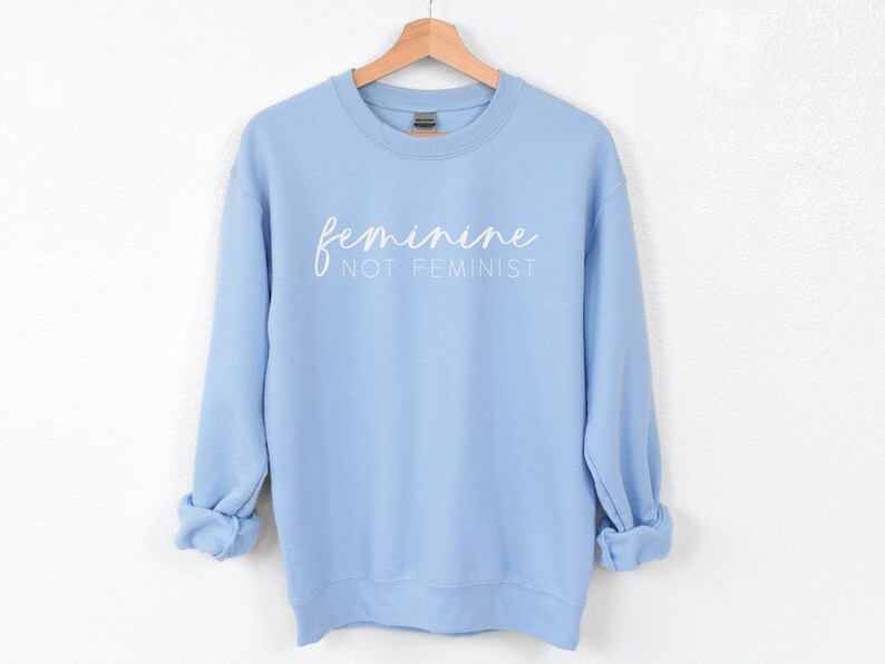 K&ouml;nnte beinhalten: Ein hellblauer Pullover mit dem Text "feminine not feminist" in wei&szlig;en Buchstaben.