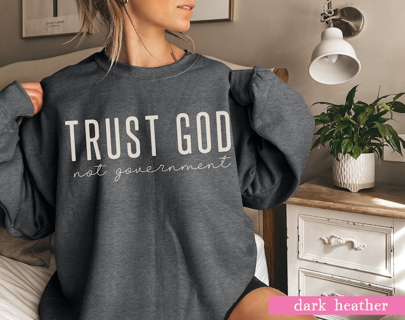 以下が含まれることがあります： 濃いグレーのヘザーのトレーナーに、白い文字で「TRUST GOD not government」と書いてあります。