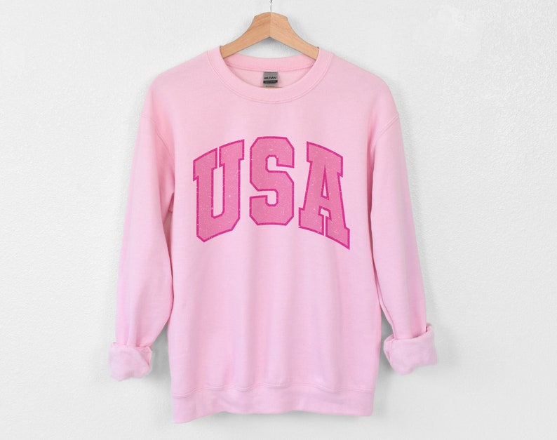 以下が含まれることがあります： 濃いピンク色の「USA」という文字がカレッジ風フォントでプリントされた、薄いピンク色のスウェットシャツ。長袖トップスは木製のハンガーに吊るされています。