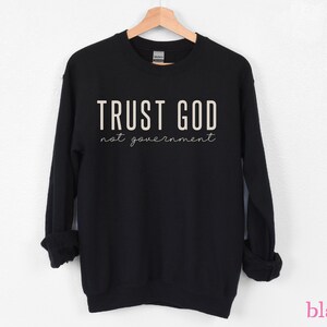 以下が含まれることがあります： 白い文字で「TRUST GOD not government」と書かれた黒いスウェットシャツ