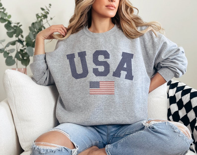 Op de afbeelding: Een grijs sweatshirt met het woord "USA" bedrukt in donkerblauwe letters. Een kleine Amerikaanse vlag is onder het woord gedrukt.
