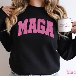 May include: Black sweatshirt with a pink "MAGA" graphic print. The sweatshirt has long sleeves and a crew neck. The model is holding a white mug with the text "Pink Patriot" in black.