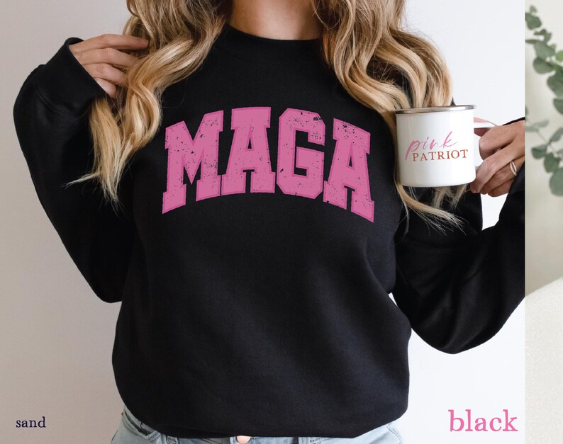 May include: Black sweatshirt with a pink "MAGA" graphic print. The sweatshirt has long sleeves and a crew neck. The model is holding a white mug with the text "Pink Patriot" in black.