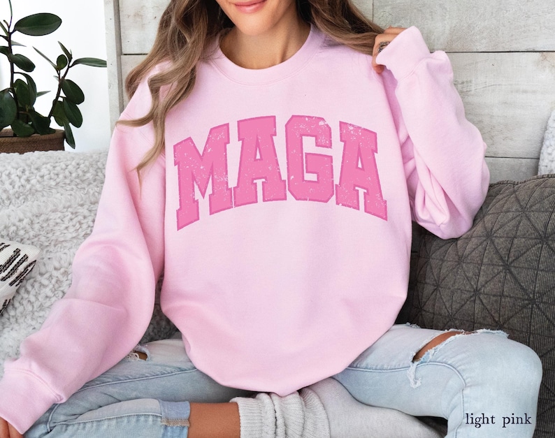 May include: A light pink sweatshirt with the text "MAGA" printed in a distressed, varsity-style font.