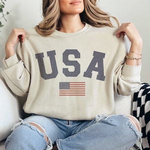 Op de afbeelding: Een beige sweatshirt met het woord "USA" gedrukt in donkergrijze letters en een kleine Amerikaanse vlag eronder. Het sweatshirt heeft lange mouwen en een ronde hals.
