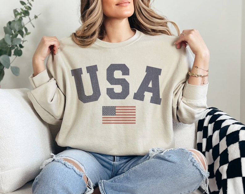 Op de afbeelding: Een beige sweatshirt met het woord "USA" gedrukt in donkergrijze letters en een kleine Amerikaanse vlag eronder. Het sweatshirt heeft lange mouwen en een ronde hals.