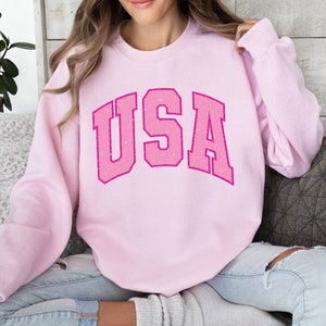 以下が含まれることがあります： 胸に「USA」の文字がかすれた明るいピンク色のスウェットシャツ。テキストはカレッジスタイルです。スウェットシャツは長袖と丸い首で、カジュアルウェアに最適です。