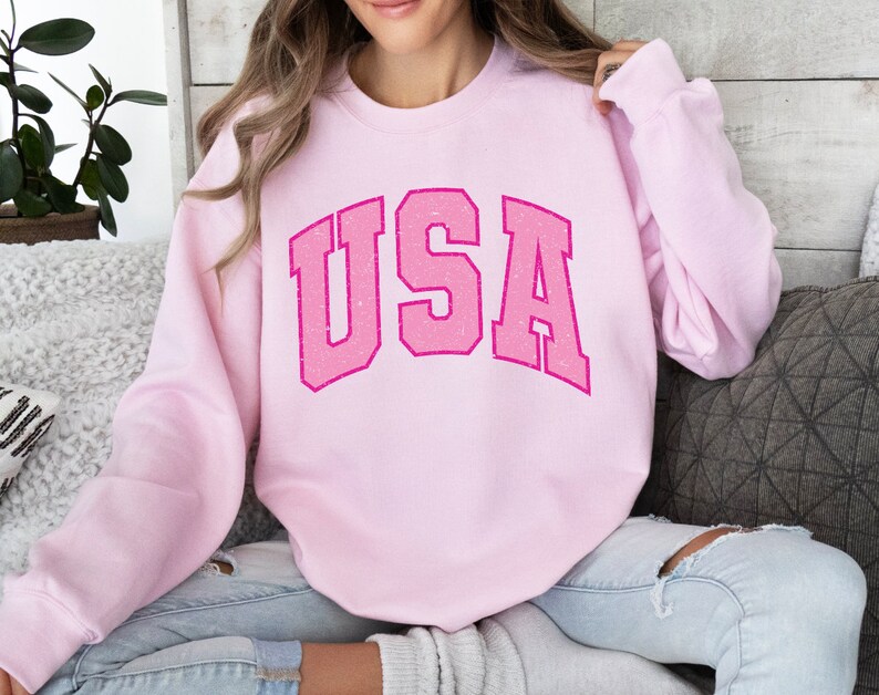 以下が含まれることがあります： 胸に「USA」の文字がかすれた明るいピンク色のスウェットシャツ。テキストはカレッジスタイルです。スウェットシャツは長袖と丸い首で、カジュアルウェアに最適です。