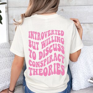 May include: A white t-shirt with a pink graphic that says "Introverted but willing to discuss conspiracy theories".