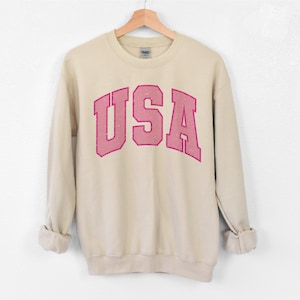 以下が含まれることがあります： ベージュのスウェットシャツ。ピンク色のブロック体で「USA」の文字がプリントされています。スウェットシャツは白い背景に木製のハンガーに掛けられています。袖は折り返されています。