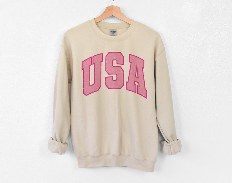 以下が含まれることがあります： ベージュのスウェットシャツ。ピンク色のブロック体で「USA」の文字がプリントされています。スウェットシャツは白い背景に木製のハンガーに掛けられています。袖は折り返されています。