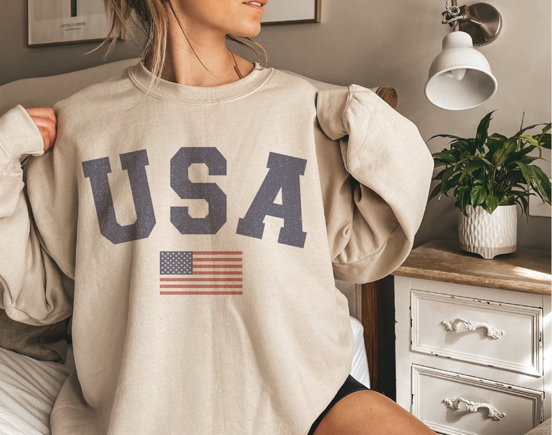 Op de afbeelding: Een beige sweatshirt met het woord "USA" bedrukt in donkerblauwe letters en een kleine Amerikaanse vlag eronder.