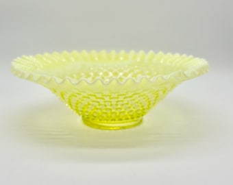 May include: A pale yellow glass bowl with a ruffled edge. The bowl has a textured, hobnail pattern on the exterior and a smooth interior. The bowl is a decorative piece, likely for serving or display.