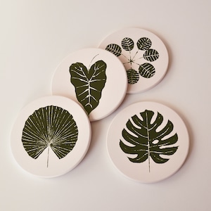 May include: Four round coasters with white backgrounds and dark green leaf designs. The coasters feature different leaf shapes, including a heart-shaped leaf, a fan-shaped leaf, and a monstera leaf.
