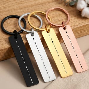 May include: Four rectangular metal keychains in black, silver, gold, and rose gold finishes. Each keychain features a circular ring and a flat, vertical pendant engraved with a unique sequence of dots and dashes representing Morse code.