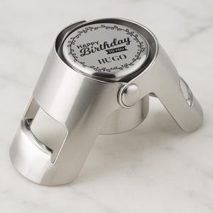 May include: A silver champagne bottle stopper with the text "Happy Birthday to You" and the name "Hugo". The stopper features a hinged design and a decorative leaf border around the top.