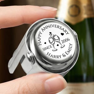 May include: A silver-coloured metal champagne bottle stopper with a circular top. The top features black engraved text that reads HAPPY ANNIVERSARY SINCE 2006 HARRY & EMILY, along with an illustration of two clinking wine glasses and small hearts.