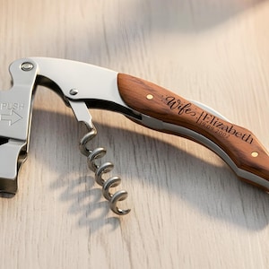 May include: A stainless steel waiter's corkscrew with a wooden handle. The metal part features a foil cutter and the word PUSH with an arrow. The wooden handle is engraved with the text Wife Elizabeth 19.09.2002 and has two small brass rivets.