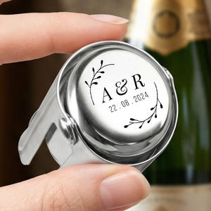May include: A silver-coloured metal bottle stopper with a circular top featuring a black engraved design of two leafy branches framing the letters A & R and the date 22.08.2024. The stopper has a smooth, polished finish and a mechanical locking mechanism.