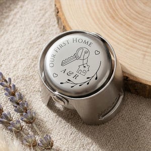 May include: A silver-toned metal bottle stopper featuring a circular top engraved with the text Our First Home, a key, a house outline, the initials A & R, and two small hearts, accented by a decorative branch design on the lower portion.