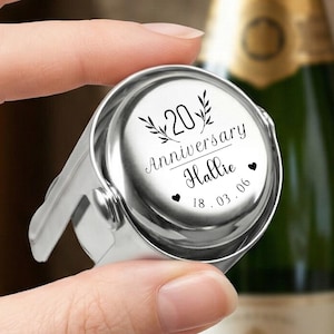 May include: A silver-coloured metal bottle stopper featuring a circular top engraved with the number 20, the word Anniversary, the name Hallie, the date 18.03.06, two small heart icons, and decorative leaf sprigs on either side of the number 20.