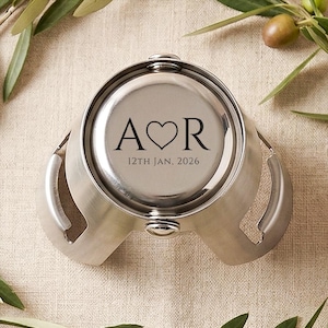 May include: A circular silver-toned metal champagne bottle stopper featuring a brushed finish. The top surface is engraved with the letters A and R separated by a heart symbol, with the date 12TH JAN, 2026 printed clearly underneath the letters.