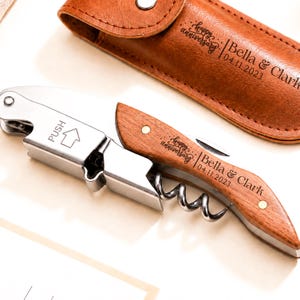 May include: A silver metal waiter's corkscrew with a wooden handle engraved with the text "happy anniversary Bella & Clark 04.11.2023" and a "PUSH" arrow icon, accompanied by a matching brown leather case with the same engraved text.